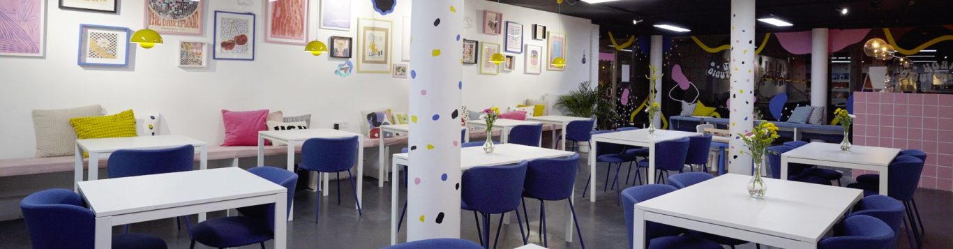 Helle Café-Atmosphäre mit weißen Tischen, bunten Stühlen und modernen Wanddekorationen.